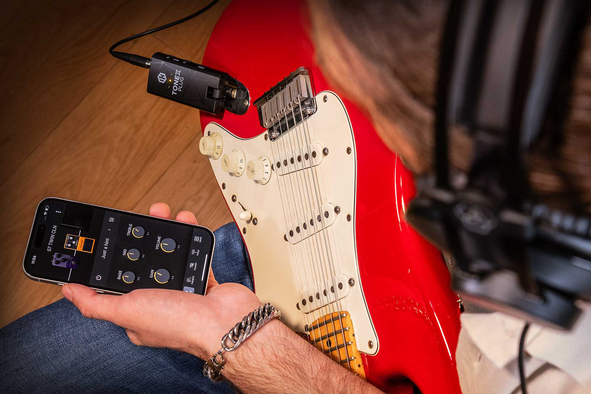 A person holds a smartphone app control screen in one hand while sitting on a red electric guitar with a Tonex headphone amplifier. The photo is taken from above, showing the person's arm, jeans, the guitar, and part of a studio setup.