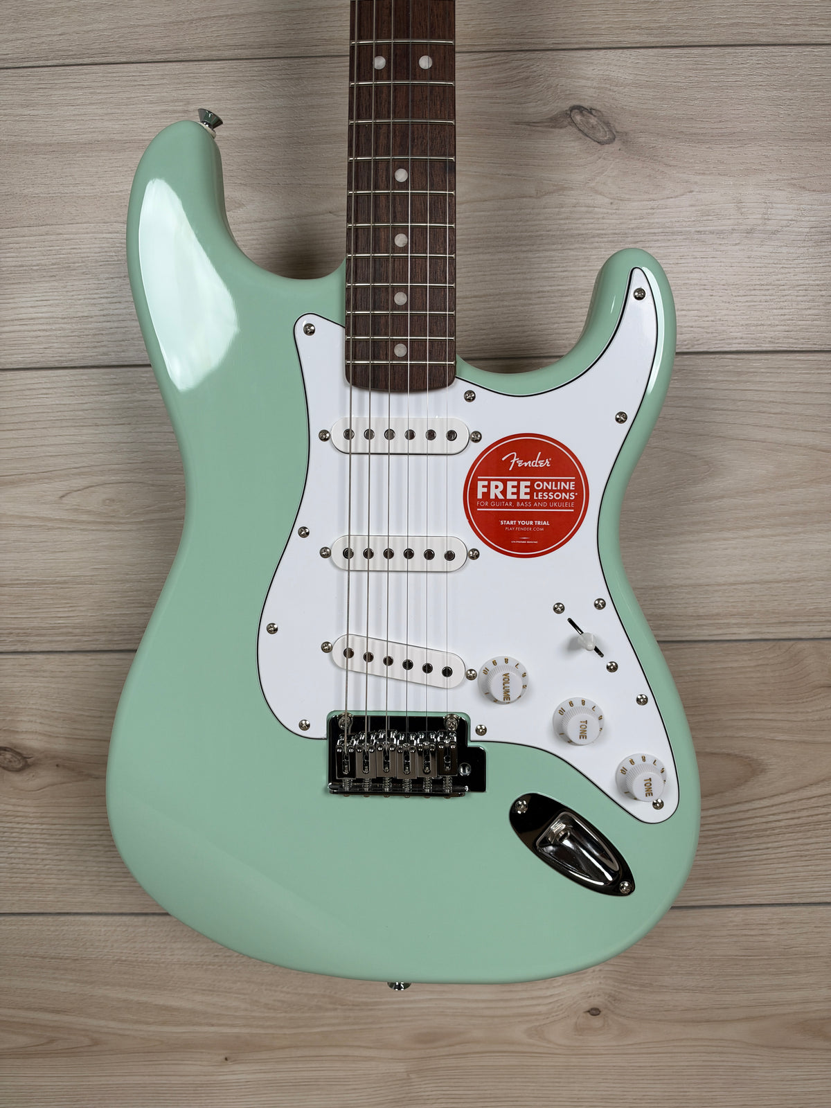 A mint green Fender Stratocaster electric guitar with a white pickguard and dark brown maple neck, laid flat on a light wood plank background.