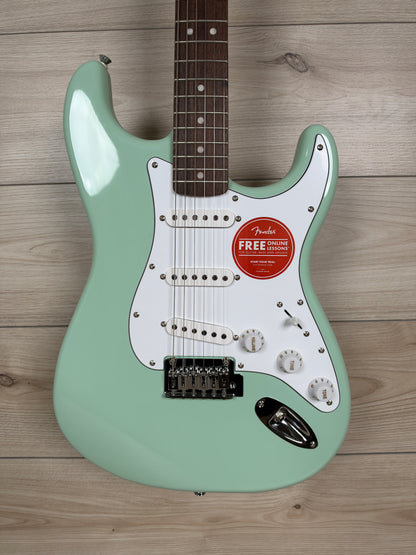 A mint green Fender Stratocaster electric guitar with a white pickguard and dark brown maple neck, laid flat on a light wood plank background.