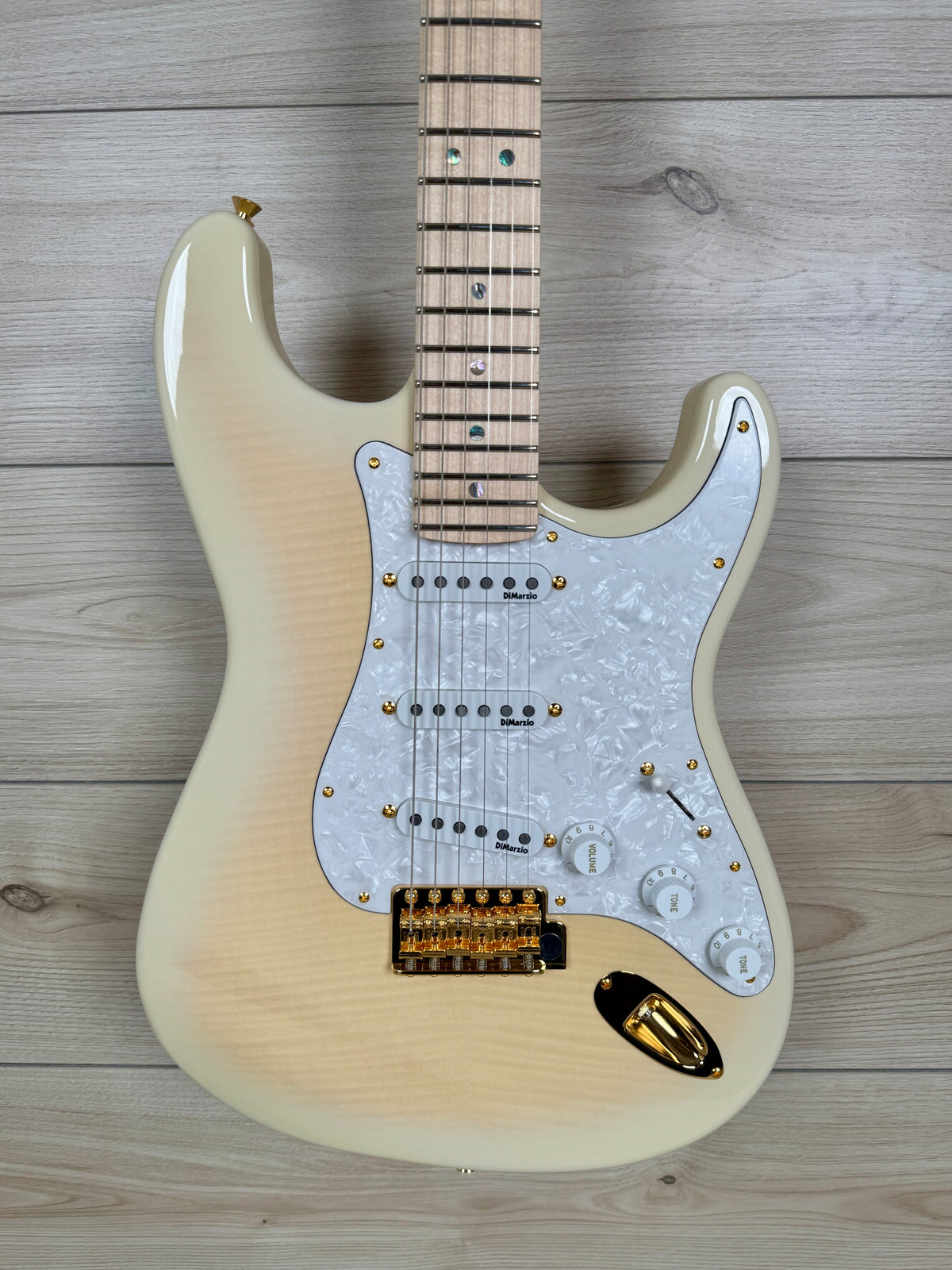 A electric guitar with a cream-colored body and a white mother-of-pearl pickguard, laid flat on a light gray wood plank background.